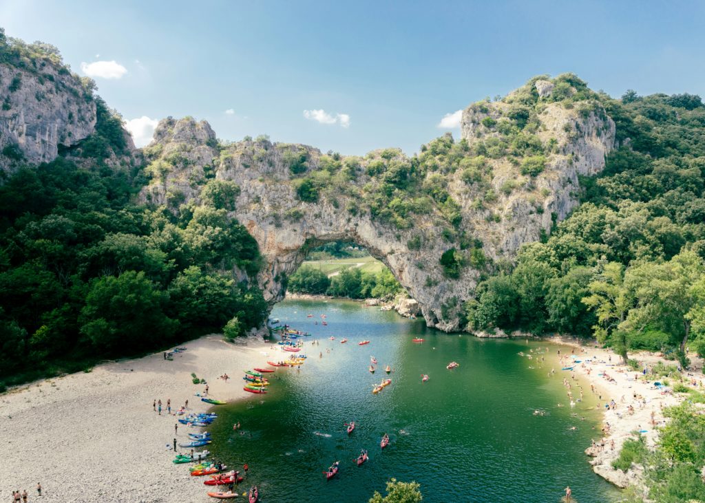 L'image représente le Pont d'Arc, arc rocheux surplombant la rivière Ardèche. Au premier plan, on aperçoit de nombreux kayaks, sur la berge de gauche et dans l'eau, tandis que la berge de droite est occupée par des baigneurs