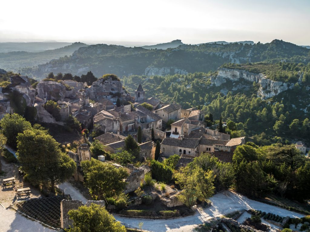 A l'avant plan de cette photo, un village provençal est perché sur la roche parmi quelques feuillus et ifs. A l'arrière plan, les monts rocheux parsemés d'arbres et bosquets se dessinent jusqu'à l'horizon